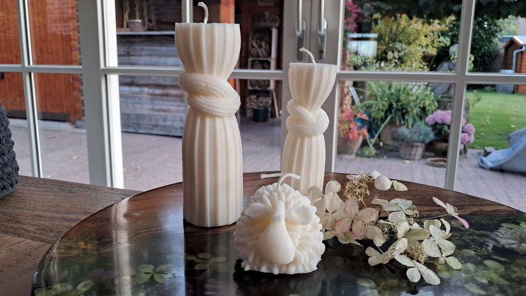 Kaars Olijk Schaap en elegante witte kaarsen op een houten tafel met een bloemdecoratie.